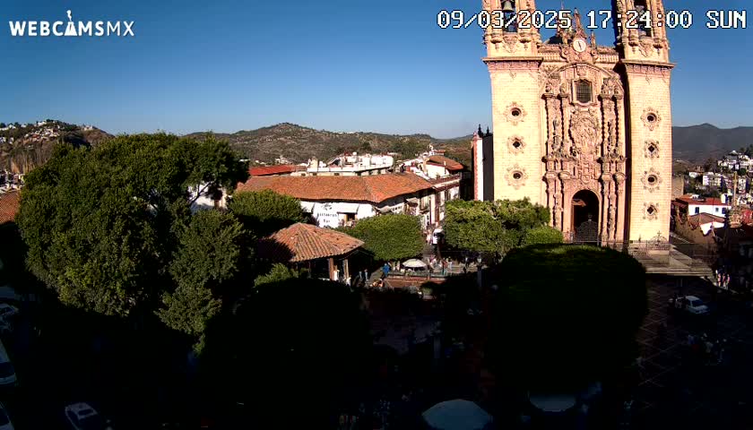 A high-angle, sunny view of a town square with a large church in the background, surrounded by buildings and trees under a clear blue sky.