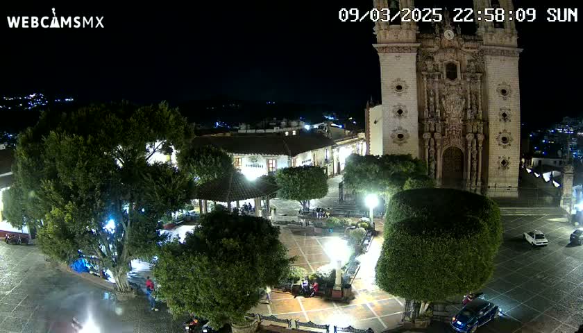 A nighttime aerial view of a town square with a large church, several cars, and lush green trees under a clear, dark sky.