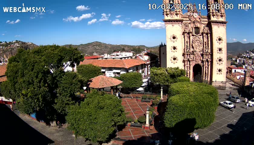 A town square with a large church, lush green trees, and a red-tiled gazebo under a sunny sky with a few scattered clouds.