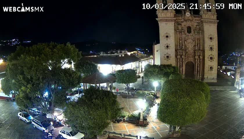 A nighttime view of a town square, showing a large church, several trees, parked cars, and a gazebo, with wet pavement indicating recent rain.