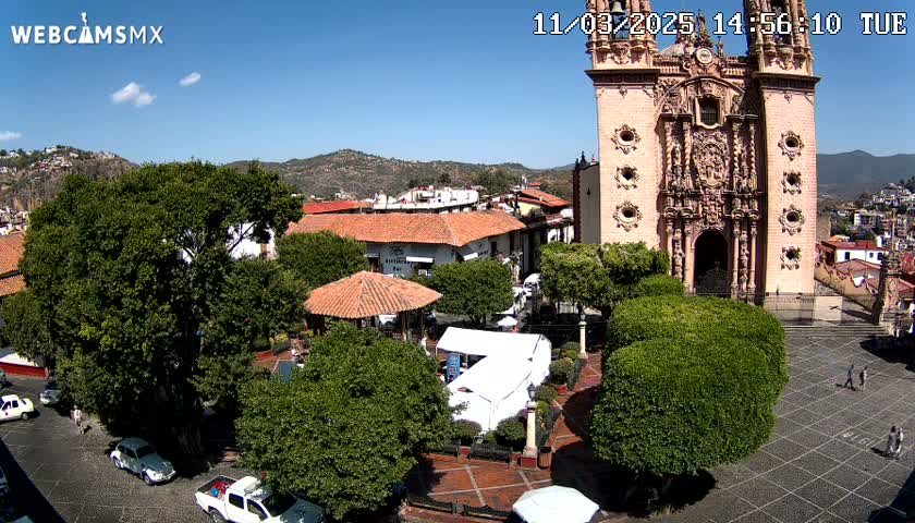A town square with a large pink church, surrounded by lush green trees and buildings under a clear blue sky.