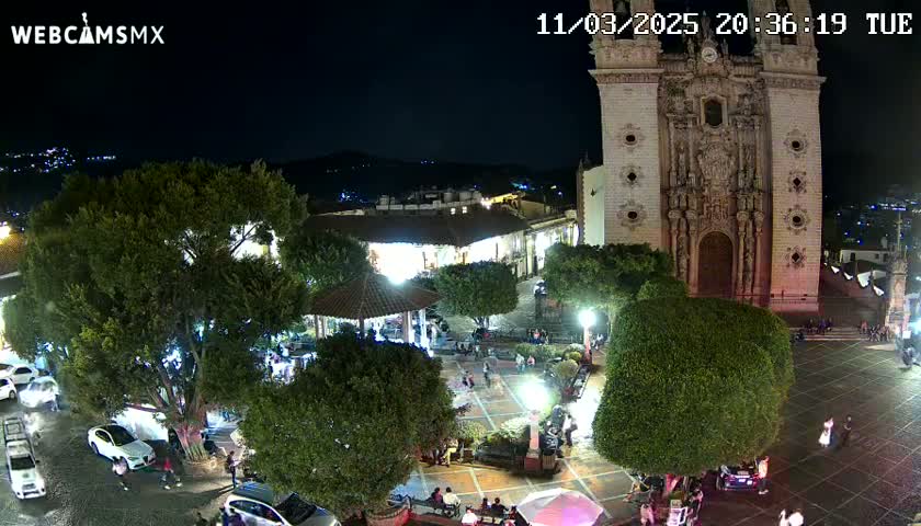 A nighttime view of a town square, illuminated, with a large church in the background and several people in the plaza.