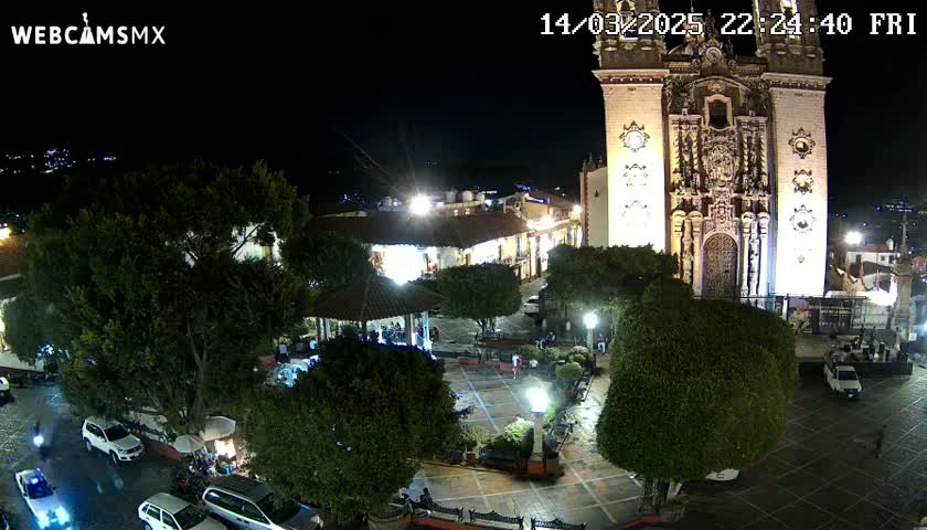 A nighttime view of a town square with a large church, illuminated buildings, trees, parked cars, and a gazebo.
