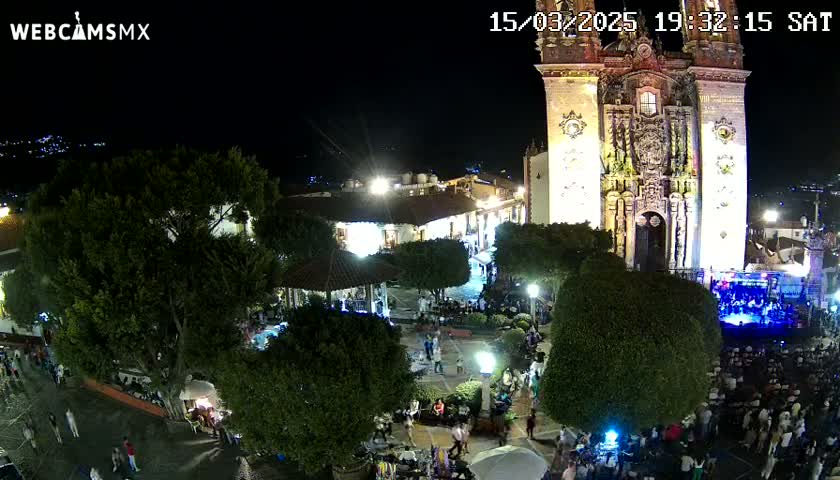 A nighttime aerial view of a town square with a large illuminated church, numerous people gathered, and lush green trees under a clear night sky.
