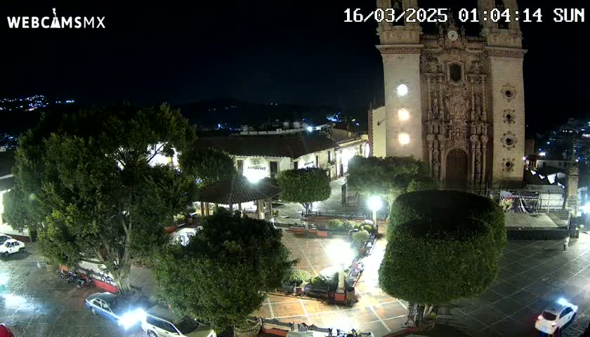 A nighttime aerial view of a town square with a large church, surrounded by trees and a few cars parked in the square.
