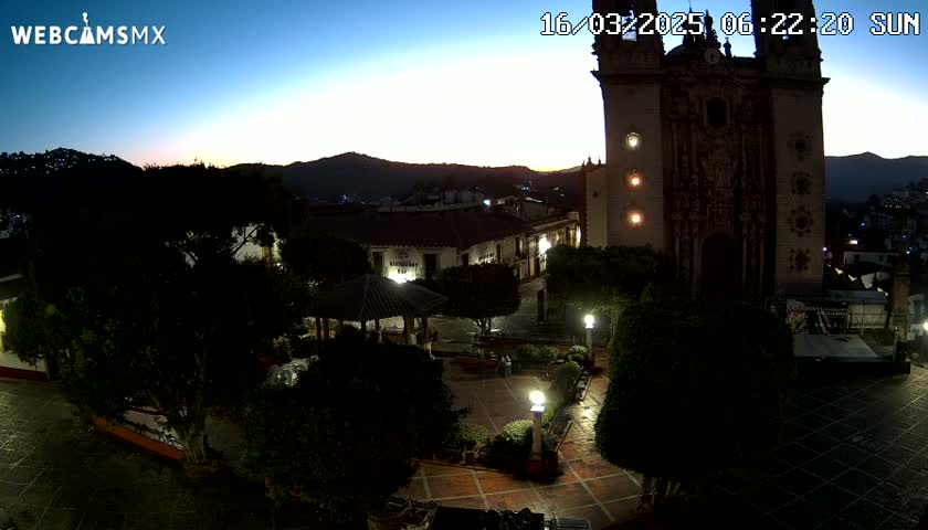A nighttime view of a town square, featuring a large church, a gazebo, and surrounding buildings under a twilight sky.