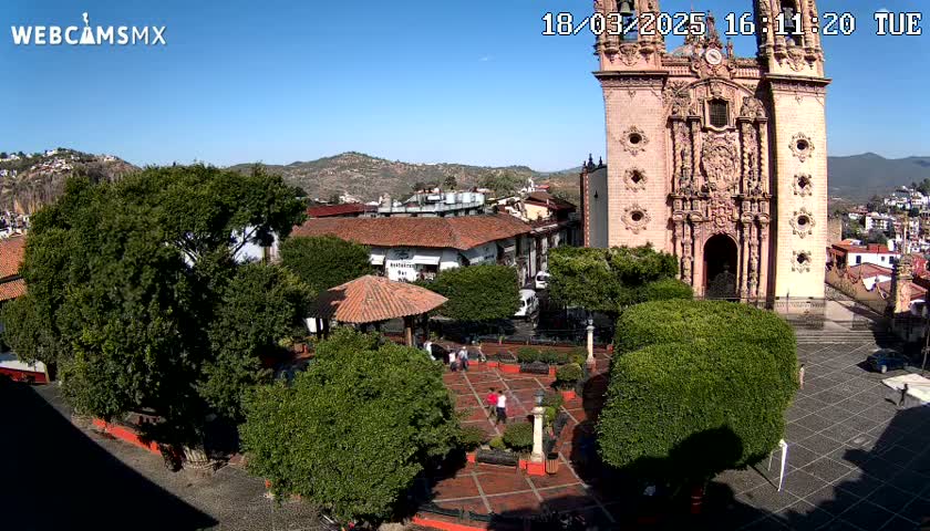 An elevated view shows a town square with a large pink church, lush green trees, and a few people under sunny skies.