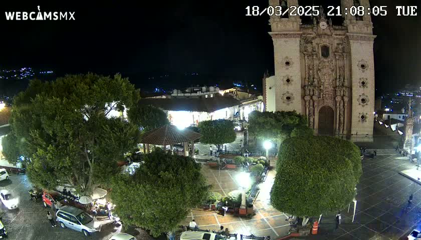 A nighttime aerial view of a town square with a large church, surrounded by trees and parked cars under clear skies.