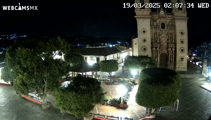 A nighttime view of a town square with a large church, several trees, and a gazebo, under a clear sky.