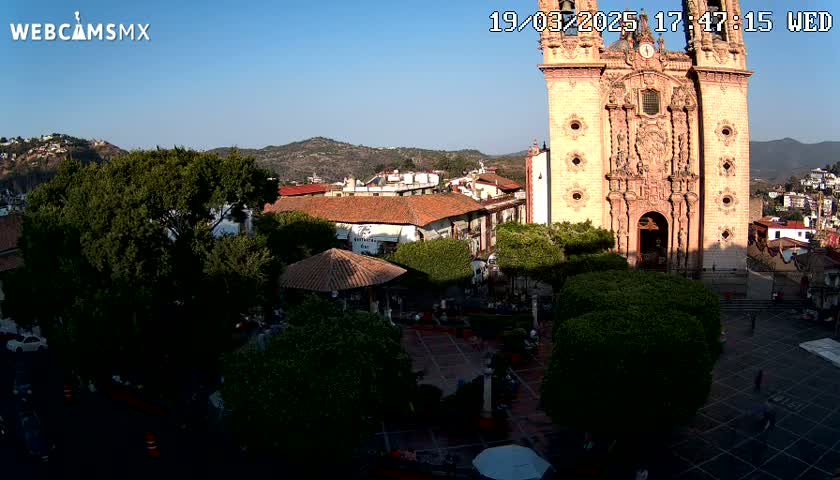 A town square with a large, ornate church in the background, under sunny skies and surrounded by lush greenery.