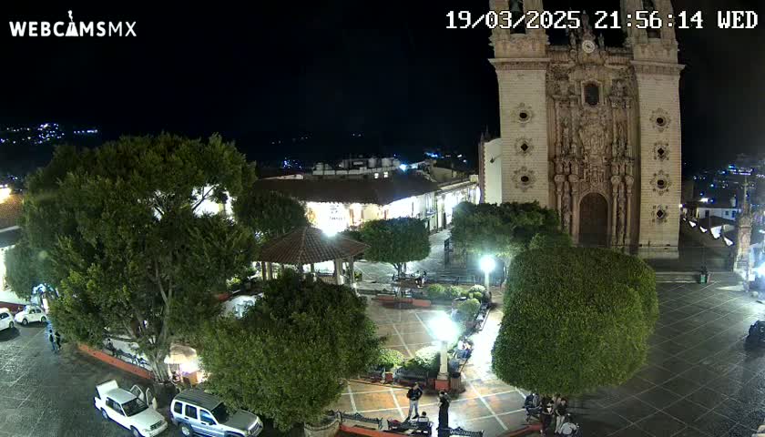 A nighttime view of a town square with a large church, several trees, a gazebo, and parked cars under a clear sky.