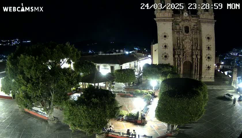 A nighttime view of a town square, featuring a large church, trees, a gazebo, and a few people, under clear night skies.