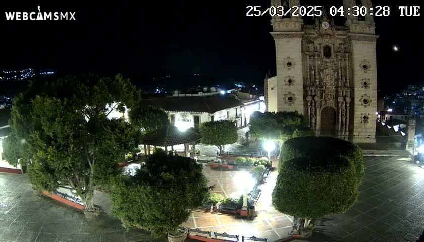 A nighttime, high-angle view of a town square featuring a large church, lush green trees, and a gazebo, all under a clear, dark sky.