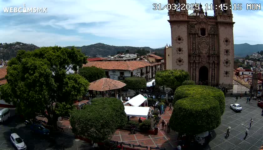 An elevated view of a town square on a partly cloudy day, featuring a large church, several buildings, lush green trees, a gazebo, and people walking around.