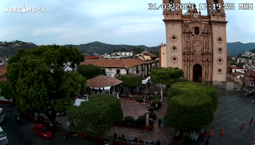 An aerial view of a town square, under a partly cloudy sky, shows a large, ornate church, a gazebo, several trees, and people gathered in the plaza.