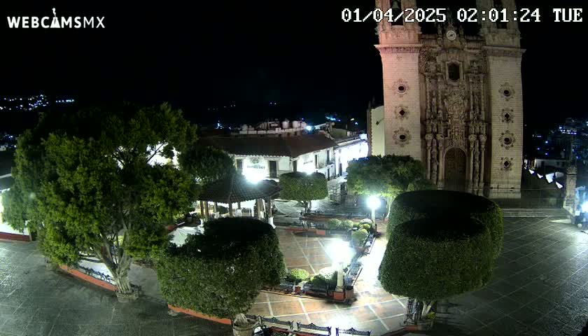 A nighttime view of a town square with a large church, illuminated trees, and a gazebo, under clear skies.