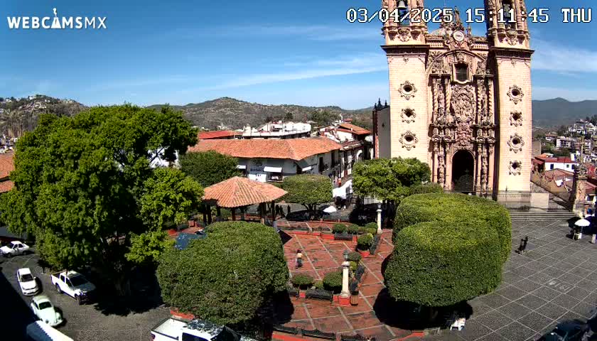 A town square with a large, ornate church in the background, under sunny skies, is surrounded by green landscaping and parked cars.
