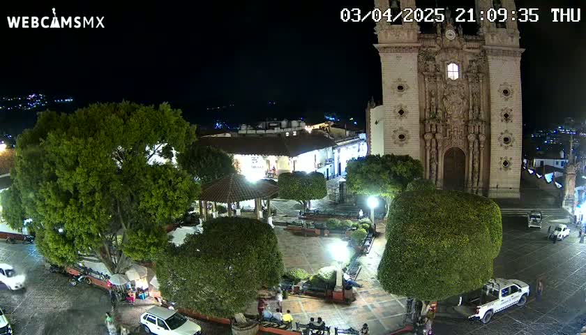 A nighttime, aerial view of a town square with a large church,  surrounded by lush green trees and shrubs, and a few parked cars and people.