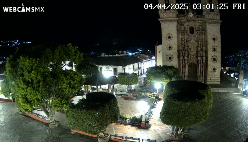 A nighttime, overhead view of a town square featuring a large church, a gazebo, lush green trees and shrubs, and paved areas, all illuminated by lights.