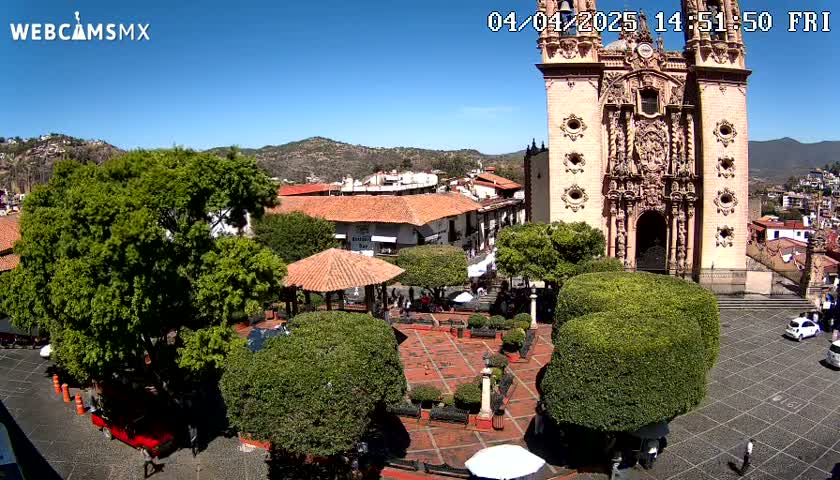 A town square with a large church, numerous green bushes and trees, and a few people, all under a clear blue sky.