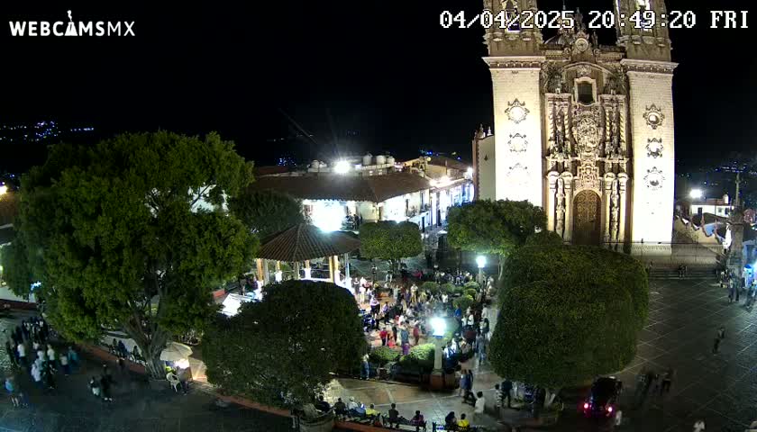 A brightly lit town square at night, filled with people, features a large church and gazebo under a clear, dark sky.
