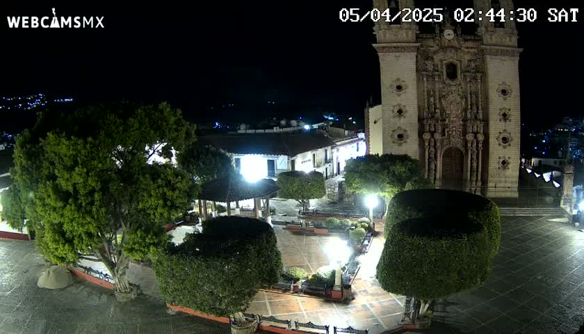 A nighttime view of a town square with a large church, several trees and shrubs, and a gazebo, under clear skies.