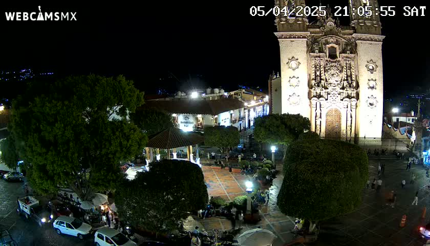 A nighttime aerial view shows a town square with a brightly lit church, gazebo, and several parked cars under a clear, dark sky.