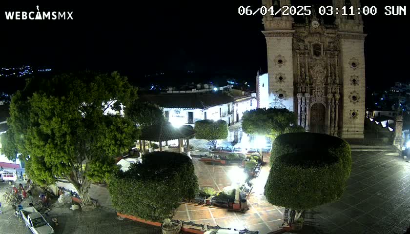 A nighttime view of a town square with a large church, several trees, and a gazebo, under clear skies.
