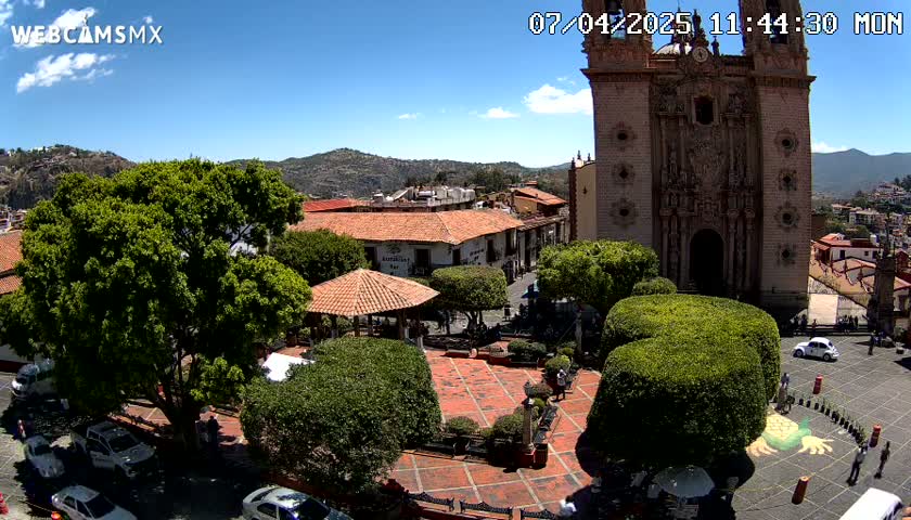 A sunny town square with a large church, several cars, and lush green landscaping, nestled in a valley against a backdrop of mountains.