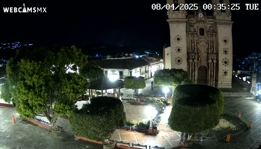 A nighttime view of a town square, featuring a large church, lush green trees, and well-lit walkways.