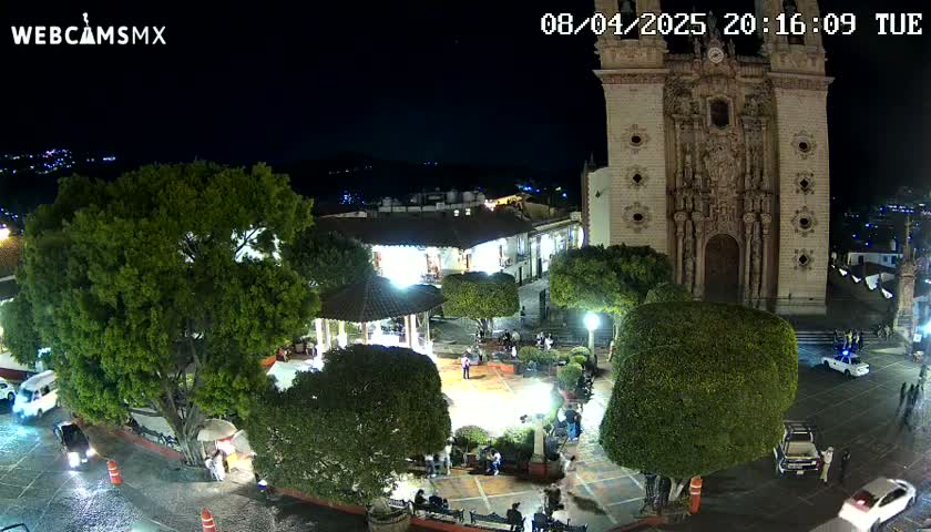 A nighttime view of a town square with a large church, lush green trees, and a gazebo, under a clear sky.