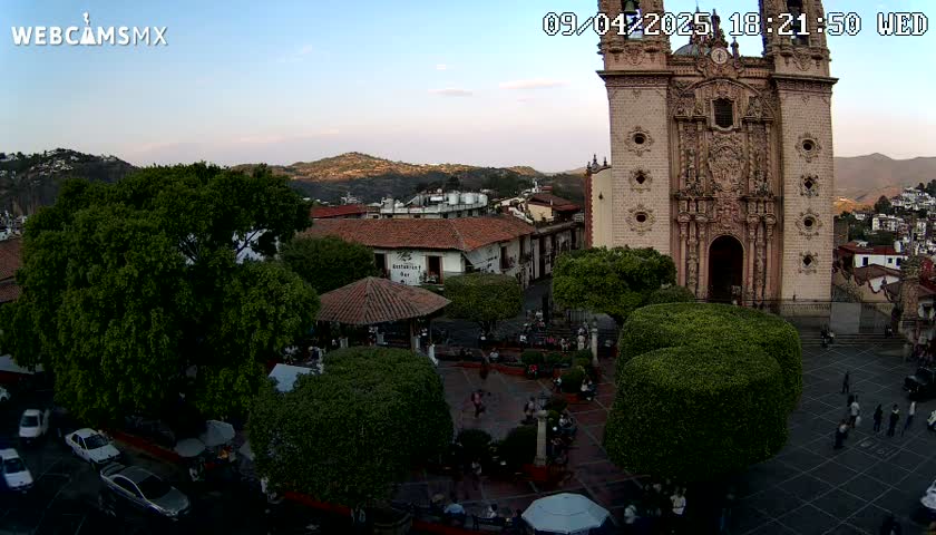A town square with a large church, numerous trees, and several people is visible under a clear evening sky.