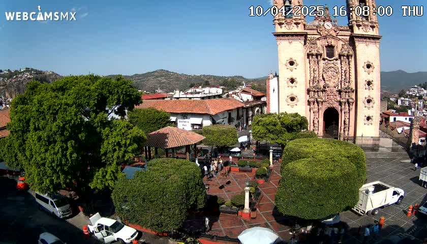 A sunny day reveals a town square centered on an ornate church, surrounded by low buildings, lush green trees, and a few people.