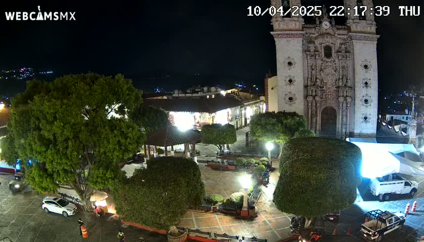 A nighttime view of a town square with a large church, surrounded by trees, landscaping, and parked vehicles under clear skies.