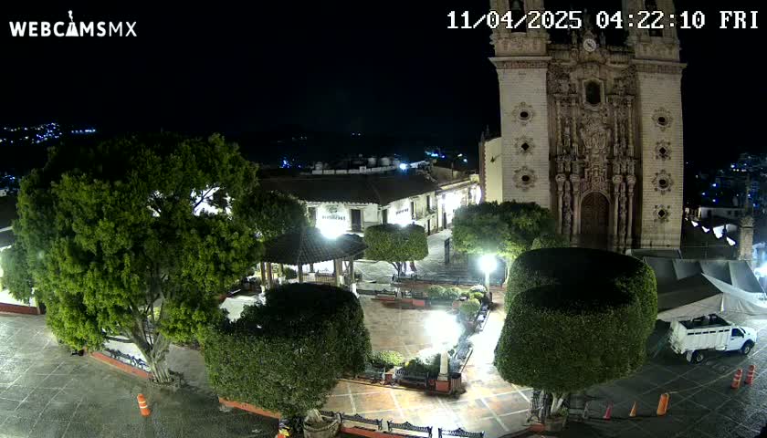 A nighttime view of a town square with a large church, lush green trees, and a gazebo, under clear skies.