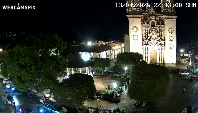 A nighttime aerial view of a town square, illuminated by streetlights, featuring a large church, a gazebo, and several vehicles parked around the perimeter, under a clear night sky.