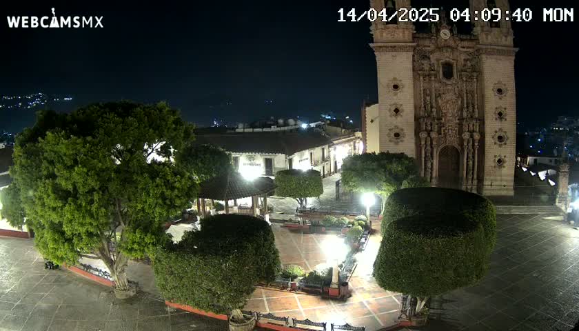 A nighttime view of a town square with a large church, trees, and a gazebo, illuminated by artificial light.