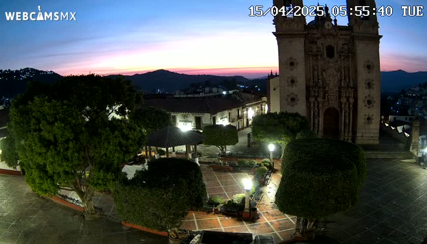 A town square at twilight, illuminated by lights, features a large church, a gazebo, and lush greenery under a mostly clear sky.