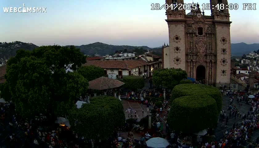 A twilight view of a town square, filled with people gathered around a plaza with a large church in the background, under a clear sky.