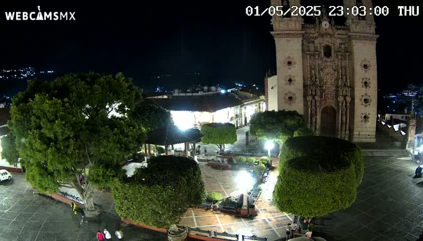 A nighttime view of a town square with a large church, numerous trees and shrubs, and a few people walking around under clear skies.