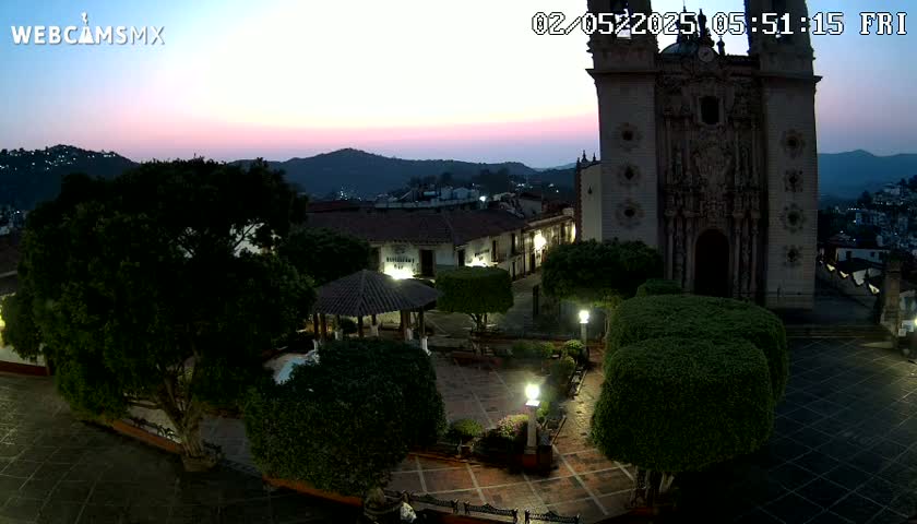 A twilight view of a town square centered around a large church, featuring low-lying shrubbery, a gazebo, and lampposts, set against a backdrop of softly lit hills.