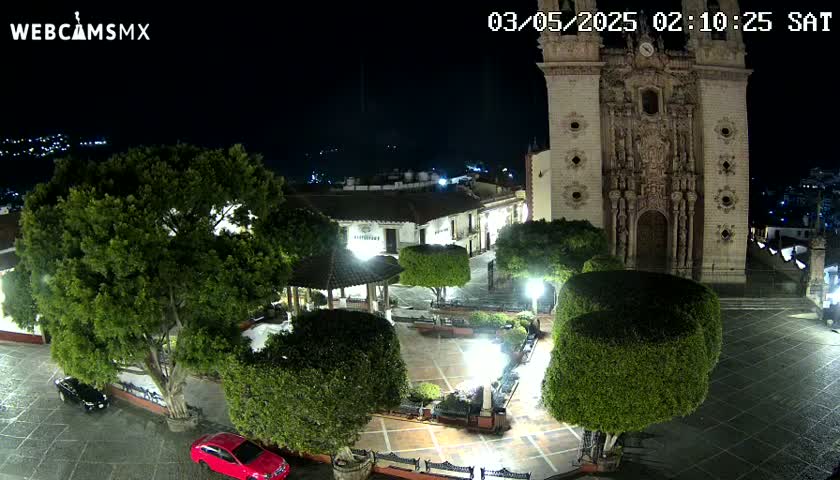 A nighttime view of a town square with a large church, several trees, a gazebo, and two cars parked.