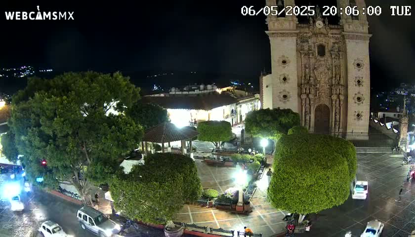 A nighttime view of a town square with a large church, lush green trees, a gazebo, and several cars on a wet street under a clear, dark sky.