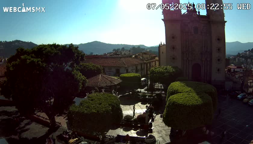 A sunlit town square with a large church, surrounded by low buildings and lush green shrubs under a clear blue sky.