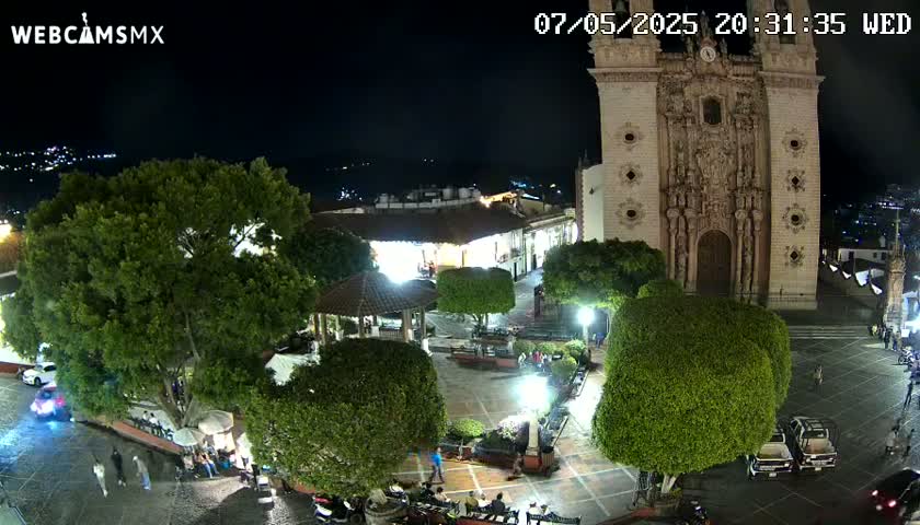 A nighttime view of a town square with a large church, several trees, and a few people walking around.