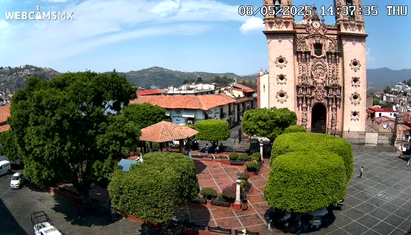 A town square with a large church, several green trees, and a gazebo is seen on a sunny day, with mountains in the background.