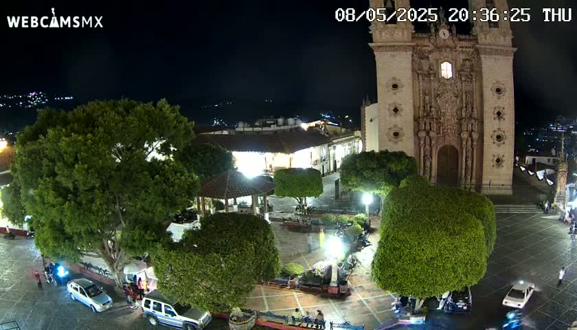 A nighttime view of a town square with a large church, several cars parked, and lush green trees under clear skies.