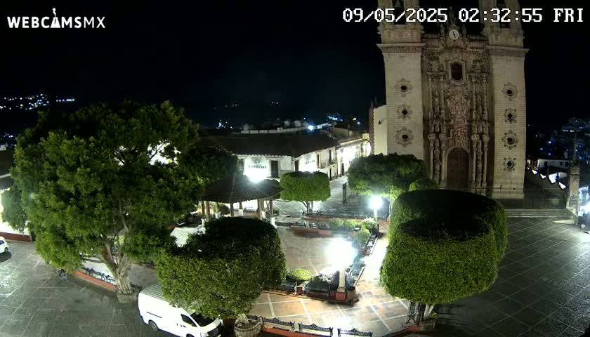 A nighttime view of a town square with a large church, numerous trees, and a gazebo, under a clear, dark sky.