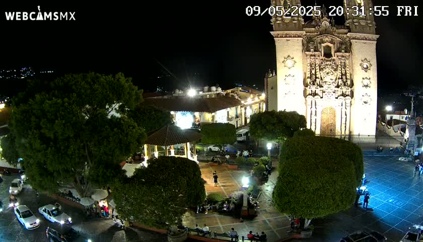 A brightly lit town square at night, featuring a large church, a gazebo, and several people, is seen under a clear, dark sky.