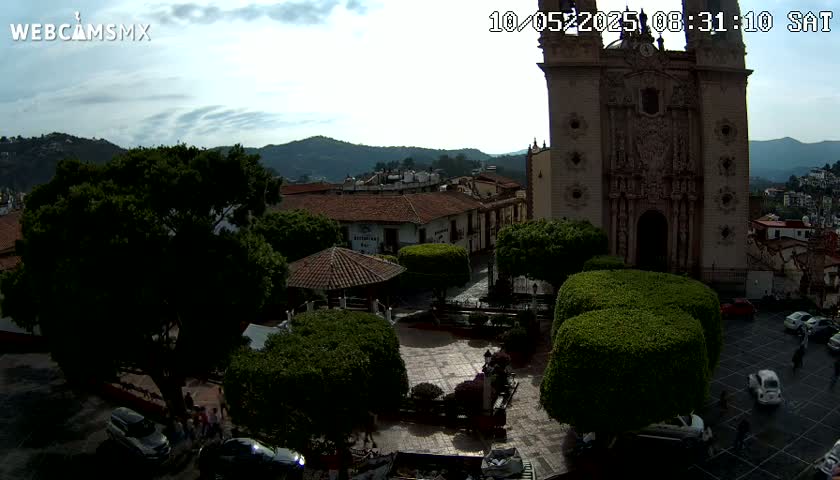 A partly cloudy day reveals a town square with a large church, several buildings, lush green shrubbery, and cars parked in a paved area, surrounded by mountains in the background.